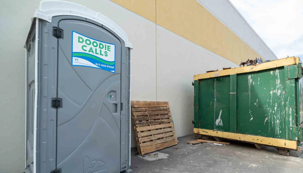 Gray ADA portable toilet unit sitting next to a building at a construction site with a green dumpster